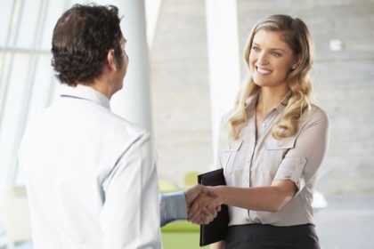 Smiling light colored woman with blond hair in a button down dress shirt holding a folder and shaking the hand of a light colored man with brown hair in a white button down shirt.