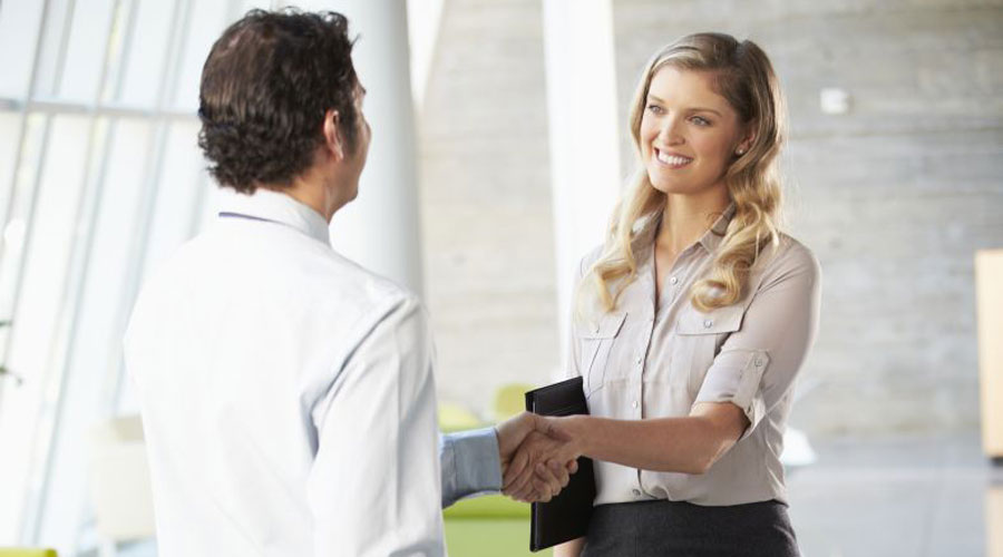 Smiling light colored woman with blond hair in a button down dress shirt holding a folder and shaking the hand of a light colored man with brown hair in a white button down shirt.