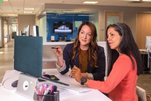 2 Females sitting at a desk in front of a computer talking.