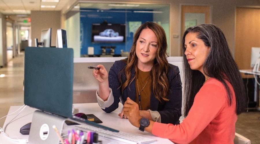 2 Females sitting at a desk in front of a computer talking.