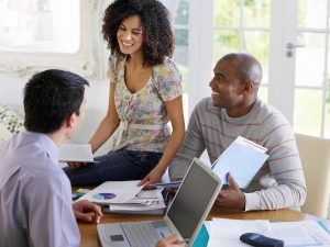 An African American female is sitting on a desk while a white male and an African American male are sitting at the desk.  There is an open laptop on the desk as well. 