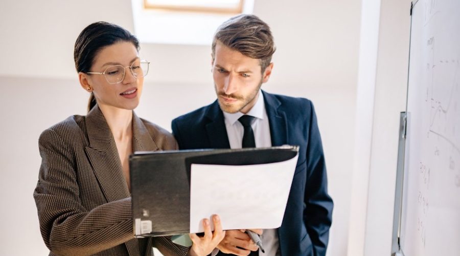 A white woman holding a notebook talking to a white male. Both are dressed in business attire