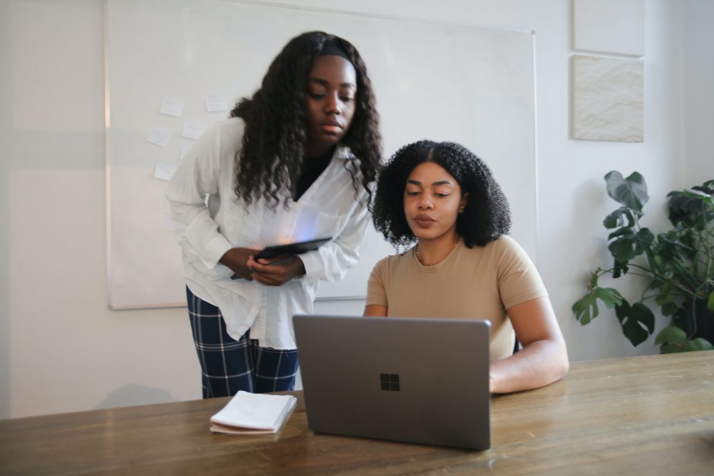 2 Woman, 1 standing and 1 sitting are talking in front of a laptop computer. 