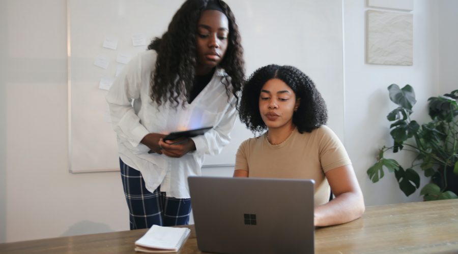 2 Woman, 1 standing and 1 sitting are talking in front of a laptop computer.