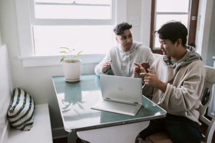 2 men are sitting in front of a laptop and are talking.