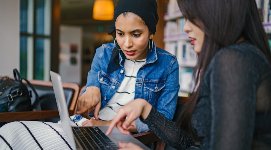 2 woman are sitting in front of a computer and talking.