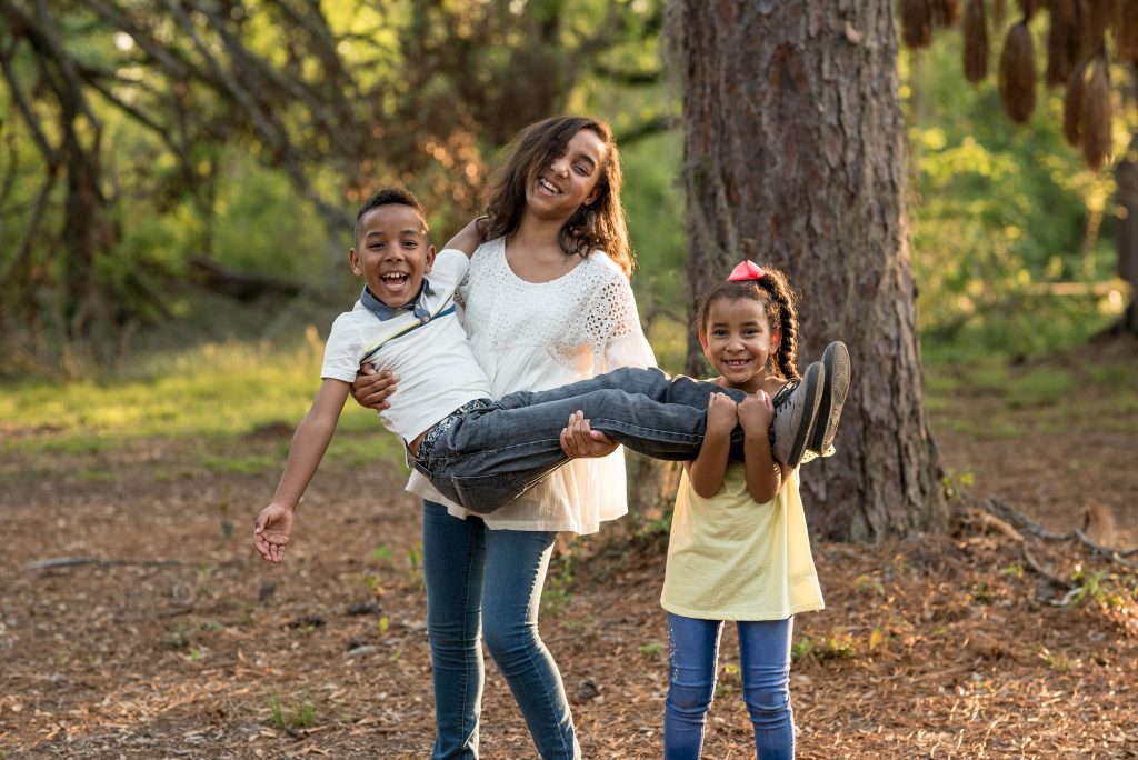 A woman is walking the park, carrying a small male child and a young girl is also with her