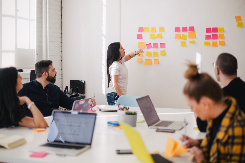 A woman is standing in front of a whiteboard and others are sitting at a conference table talking with her