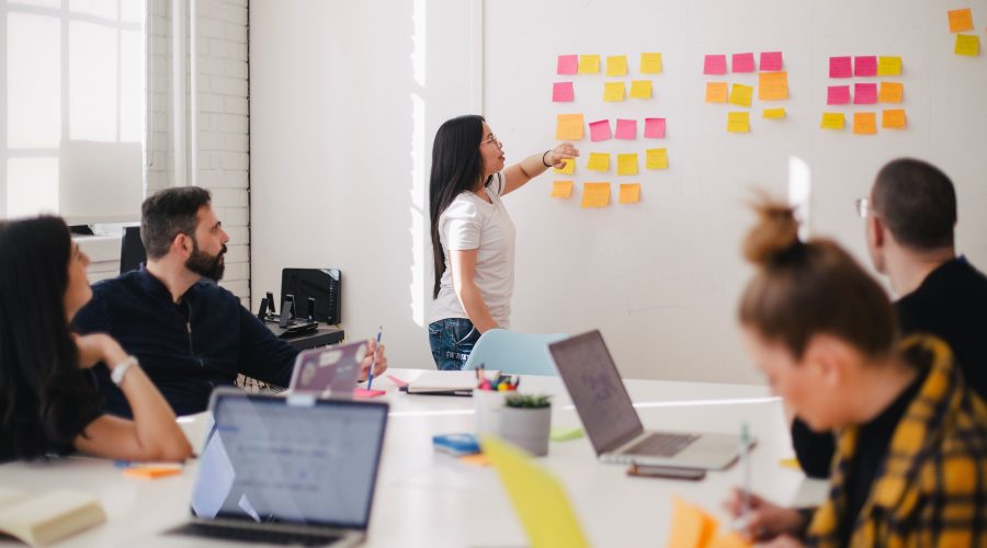 A woman is standing in front of a whiteboard and others are sitting at a conference table talking with her