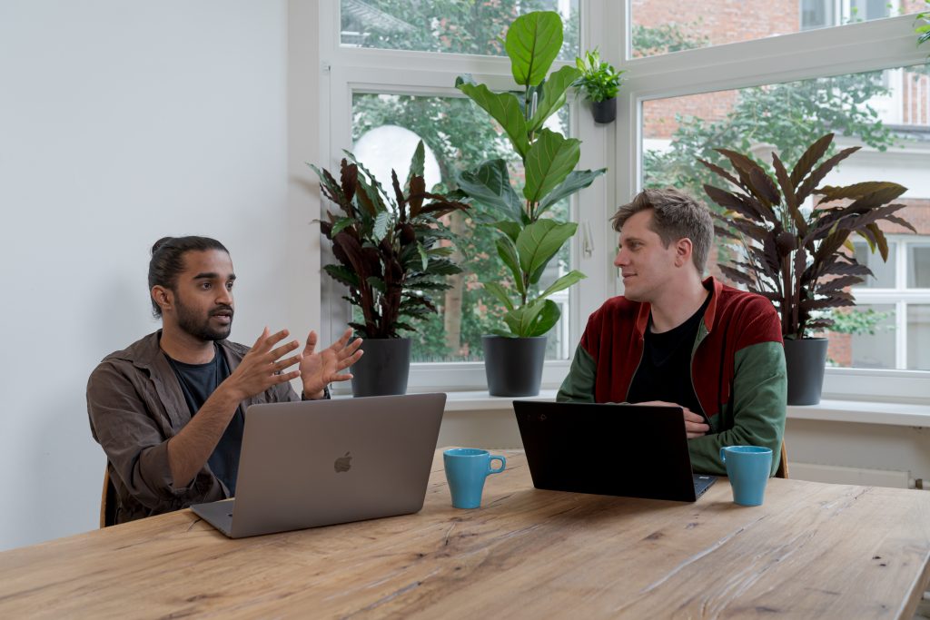 Two men are sitting at a table with computers and talking