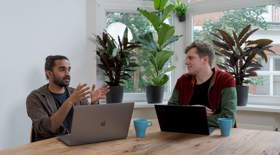 Two men are sitting at a table with computers and talking