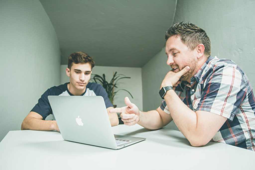 2 Men are sitting at a table talking in front of an open laptop computer