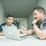 2 Men are sitting at a table talking in front of an open laptop computer