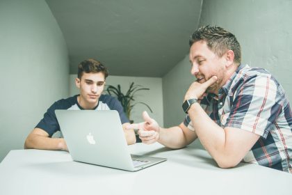 2 Men are sitting at a table talking in front of an open laptop computer