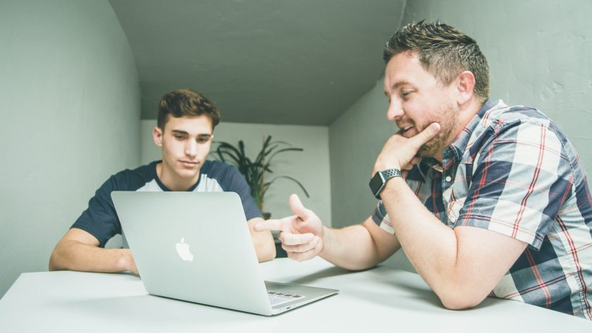 2 Men are sitting at a table talking in front of an open laptop computer