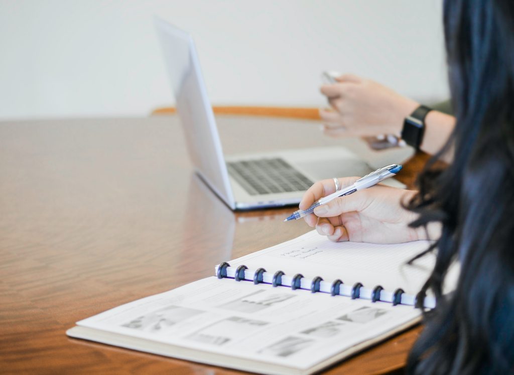 Someone is sitting at a desk with a notepad and pen, and a laptop computer is open 