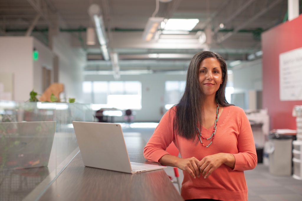 A business woman is standing at a table with her laptop computer open.