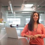 A business woman is standing at a table with her laptop computer open.