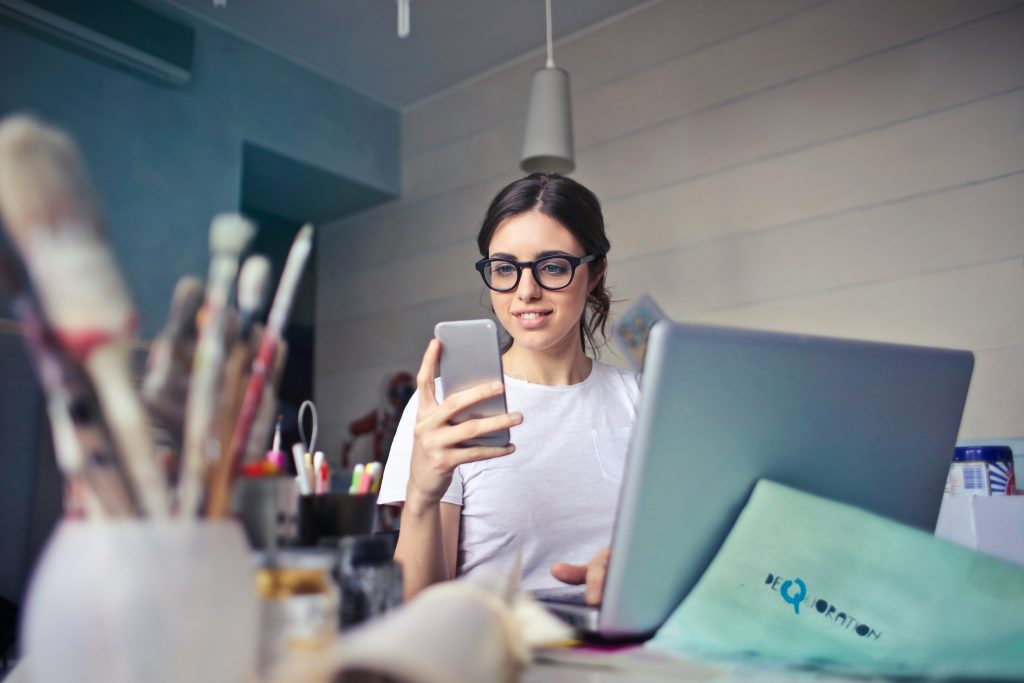 A woman is sitting in front of laptop computer and is also holding a smartphone. 
