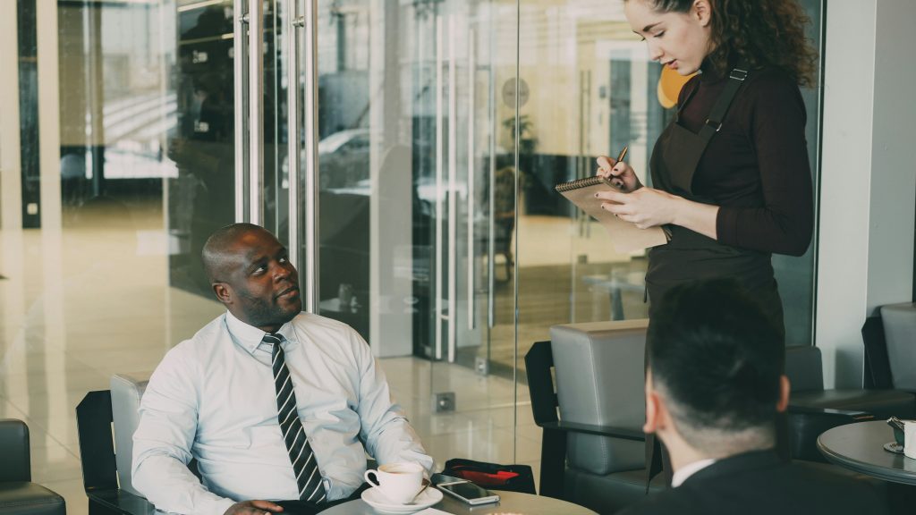 2 Business men are sitting while a woman sit standing and writing down notes