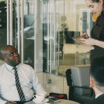 2 Business men are sitting while a woman sit standing and writing down notes