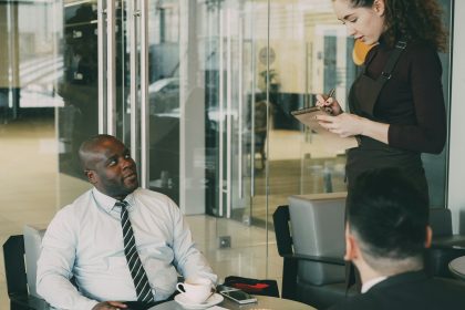 2 Business men are sitting while a woman sit standing and writing down notes