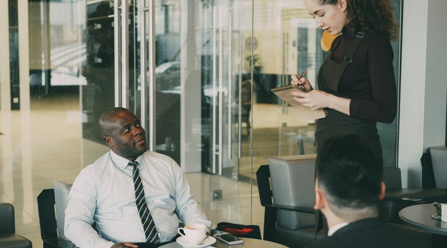 2 Business men are sitting while a woman sit standing and writing down notes
